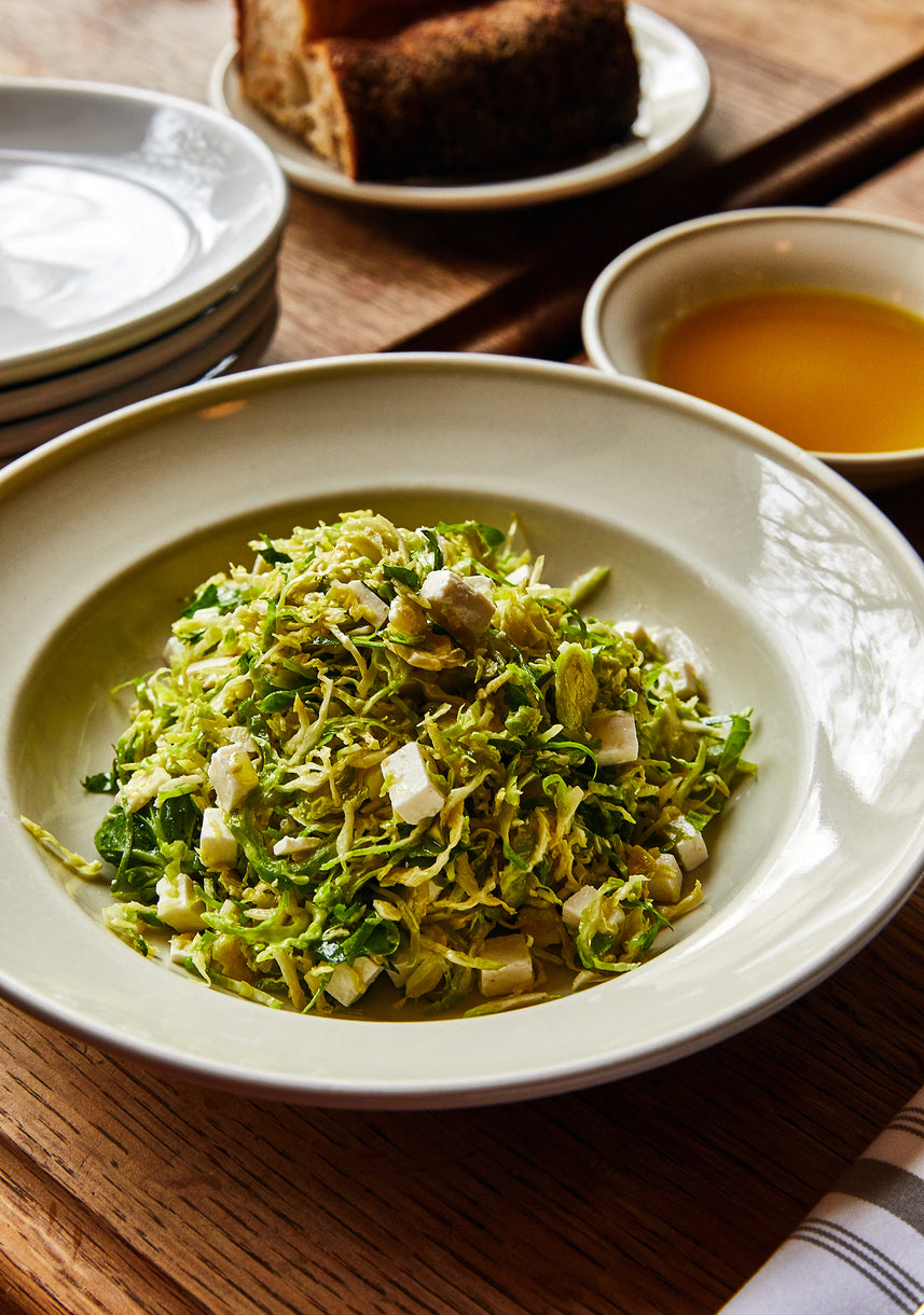 A shallow white bowl filled with a fresh shaved Brussels sprouts salad mixed with small cubes of cheese. In the background, part of a small bowl with golden olive oil is visible, all set on a wooden table
