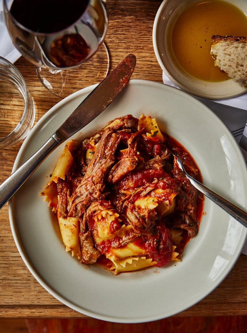 A plate of wide ribbon pasta topped with shredded meat and rich tomato sauce, served with a knife and spoon. In the background, there is a glass of red wine, a bowl of olive oil with a piece of bread, and part of a water glass, all set on a wooden table