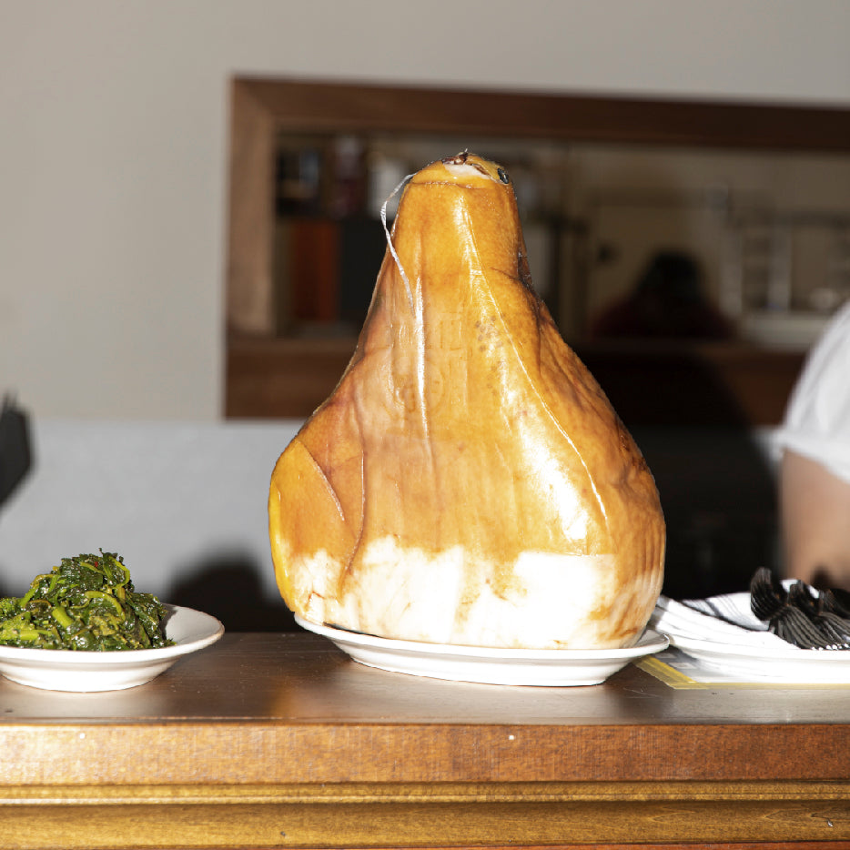 A large cured ham, likely prosciutto, displayed upright on a plate, next to a smaller plate of cooked leafy greens, set on a wooden counter with a blurred background