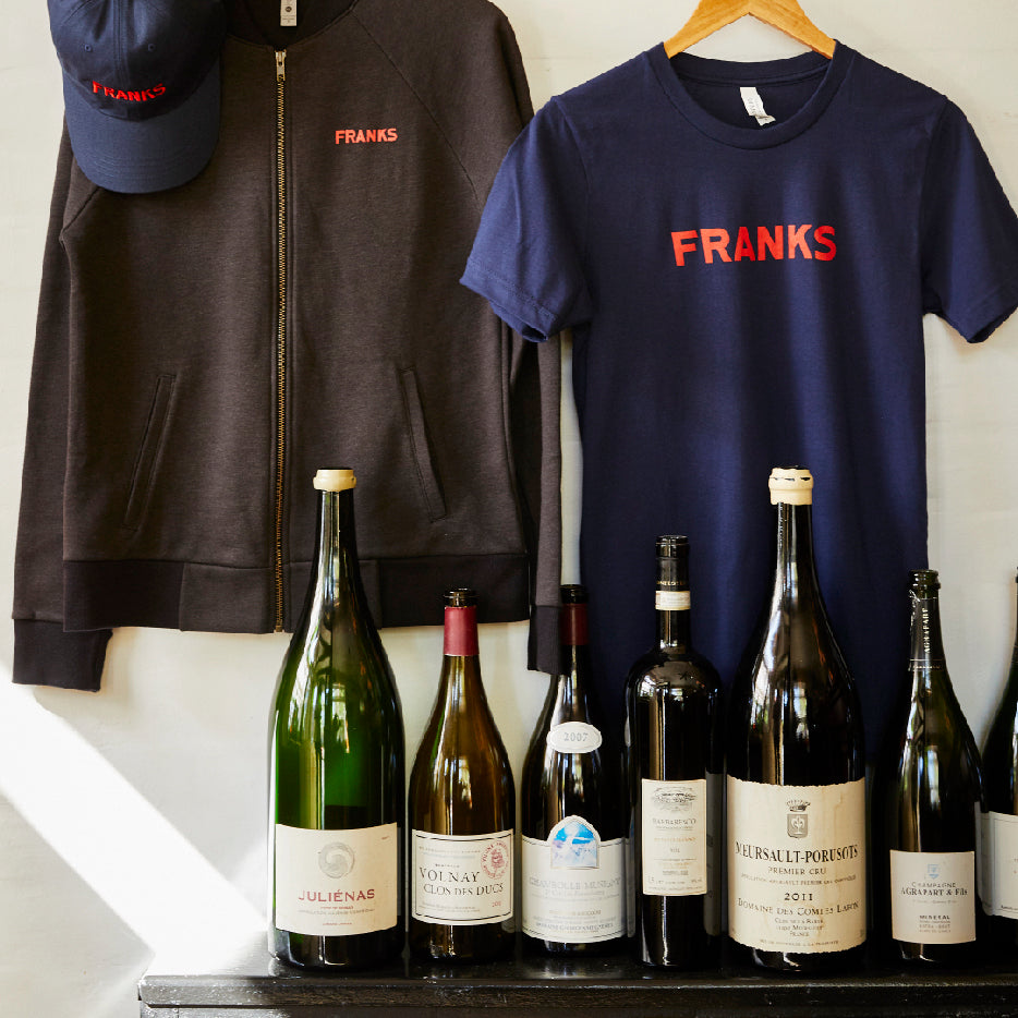 A display of FRANKS merchandise and wine bottles. On the left, a dark zip-up hoodie with a red "FRANKS" logo is paired with a matching cap. On the right, a navy blue T-shirt with the same red "FRANKS" logo hangs on a wooden hanger. In front, an assortment of wine bottles, including magnums and standard sizes, are arranged in a row, showcasing various wine labels
