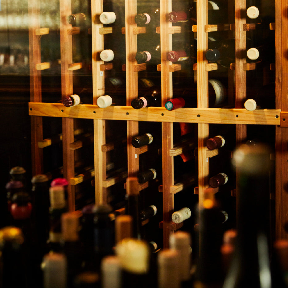 A wooden wine rack filled with bottles stored horizontally, organized with necks facing outward. The rack is divided into sections with light-colored wooden supports. In the foreground, several wine bottles are slightly out of focus, adding depth to the warm, softly lit scene