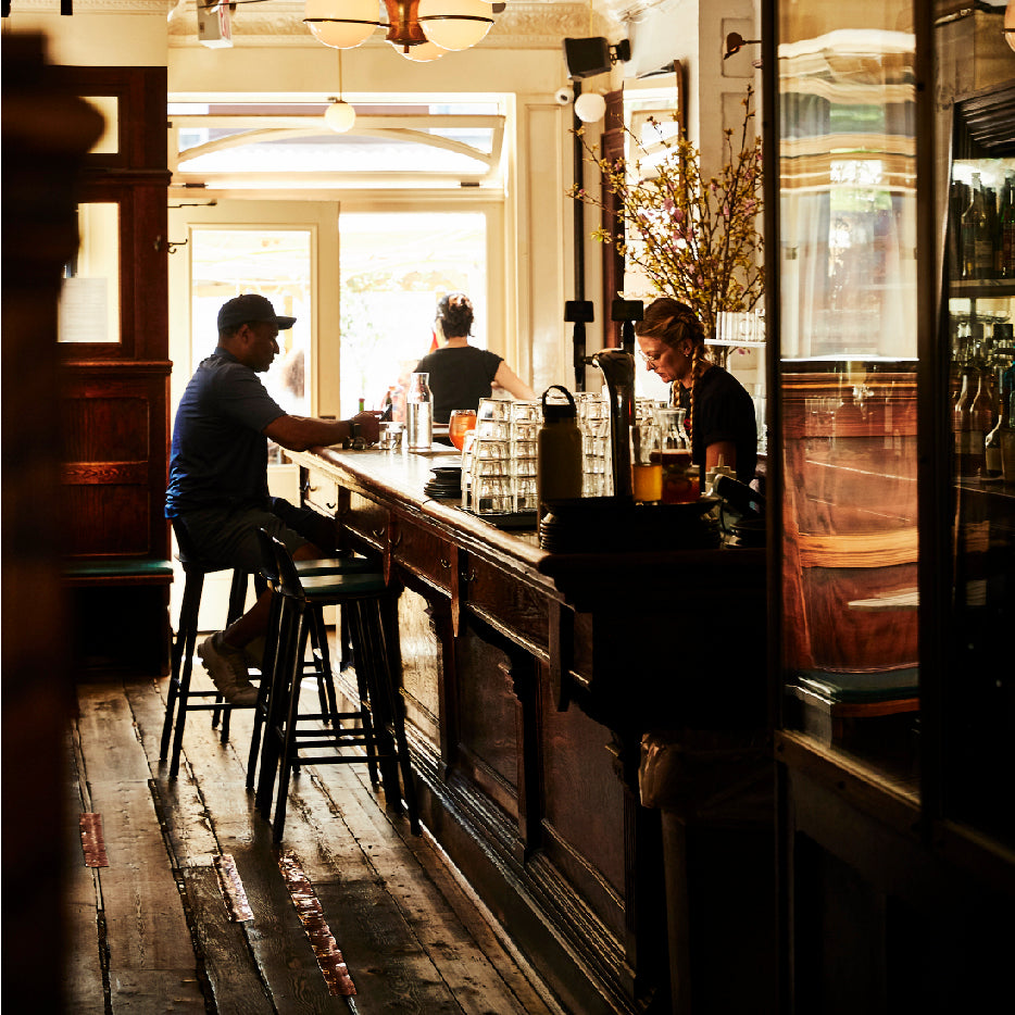 A warmly lit bar interior with wooden floors and dark wood paneling. A bartender serves drinks to a man wearing a cap seated on a high stool at the counter, while another person stands near the entrance with sunlight streaming through the glass door. Glassware, bottles, and vases with flowers are arranged along the bar, creating a cozy and inviting atmosphere