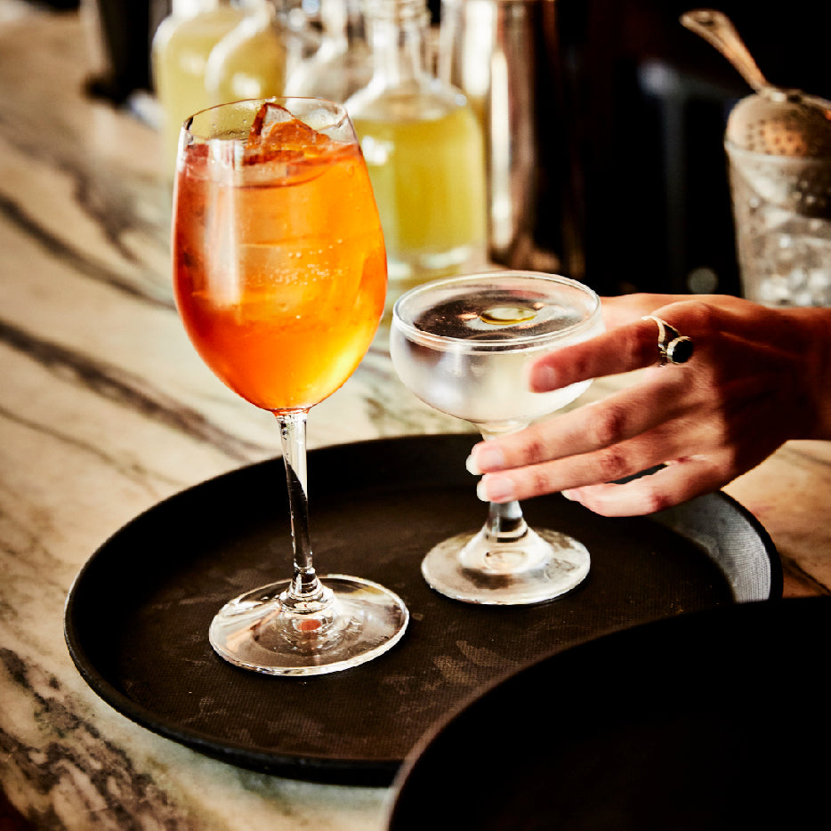 A close-up of two cocktails on a black serving tray placed on a marble bar counter. One drink is an orange Aperol Spritz in a wine glass with ice, and the other is a clear cocktail in a coupe glass garnished with an olive, being picked up by a hand. In the background, bottles filled with light yellow liquid and a cocktail strainer are visible, giving the scene a sophisticated bar atmosphere.