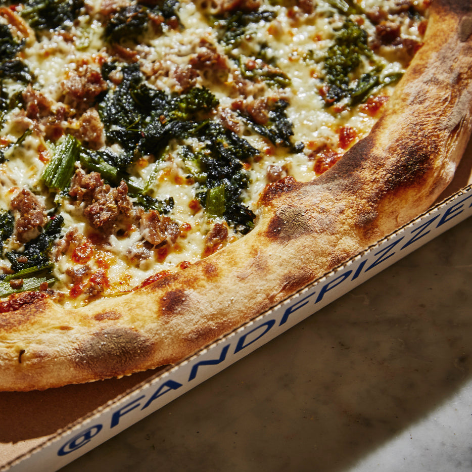 Close-up of a pizza with a golden, blistered crust, topped with melted cheese, crumbled sausage, and sautéed broccoli rabe, presented in an open pizza box
