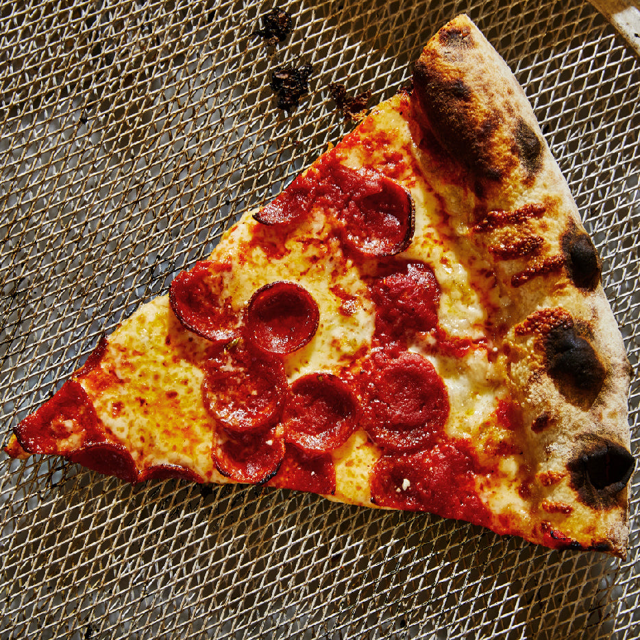A slice of pepperoni pizza with a bubbly, charred crust, melted cheese, and crisped pepperoni slices, resting on a mesh pizza screen