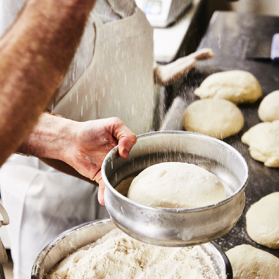 A close-up of a baker dusting flour over a metal bowl containing a round ball of pizza dough. Below, another container is filled with flour, and in the background, several dough balls are arranged on a work surface, ready for baking. The scene captures a hands-on moment in the pizza-making process
