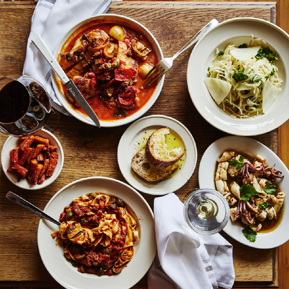 An overhead view of a wooden table set with a variety of Italian dishes, including a plate of braised meat in tomato sauce with potatoes, a pasta dish with shaved cheese and parsley, a seafood salad with calamari, a bowl of tagliatelle in a rich tomato sauce, a small plate of roasted carrots, and a plate with sliced bread served alongside olive oil. Glasses of red and white wine accompany the meal, along with white napkins.