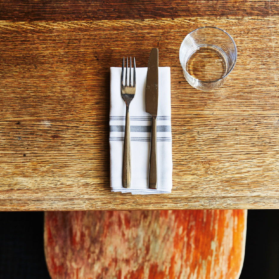 A fork and knife placed on a napkin on a wooden table, accompanied by a glass, viewed from above.