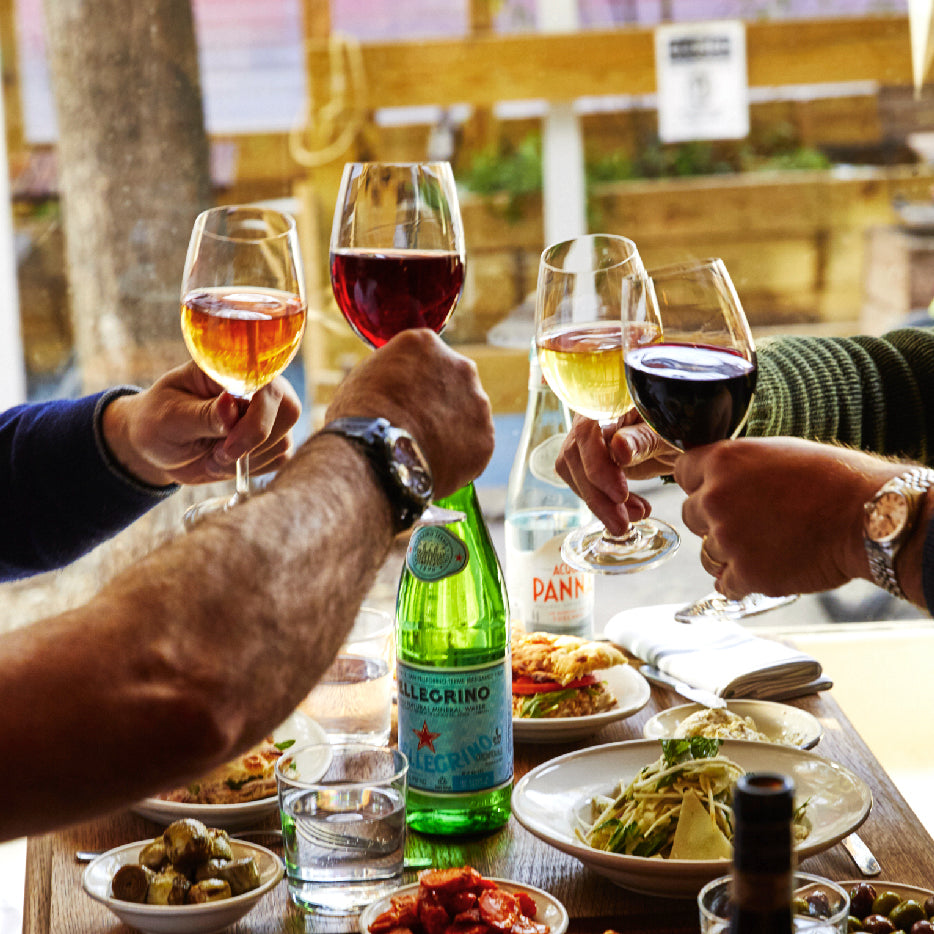 Four people clink wine glasses over a table filled with Italian dishes, including pasta, olives, and small plates of appetizers. A bottle of San Pellegrino sparkling water and an Acqua Panna still water bottle are at the center of the table. The scene is set in a bright, casual dining environment with natural light streaming in from the window.