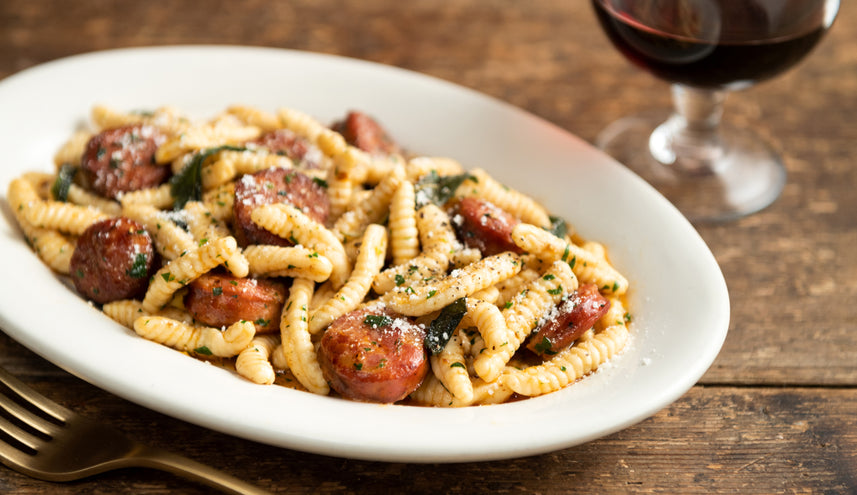 A white oval plate filled with twisted pasta tossed with slices of browned sausage, fresh herbs, and grated cheese, served on a rustic wooden table with a fork nearby and a glass of red wine in the background