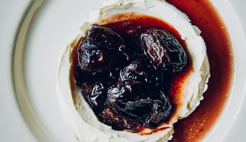 A wheel of creamy cheese topped with glossy, dark poached or preserved fruits in a rich syrup, served on a white plate with the syrup pooling around the edges