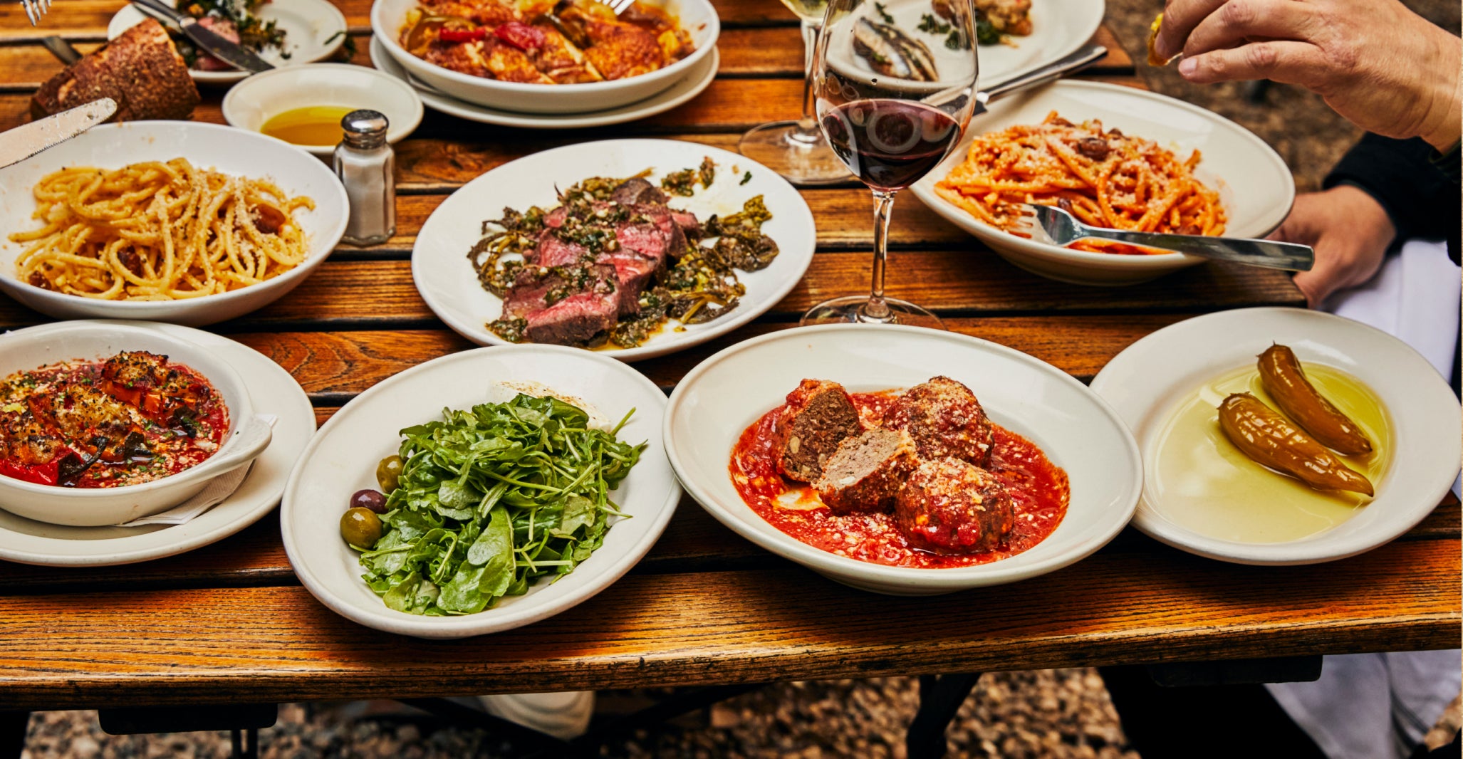 A rustic wooden table set with an assortment of Italian dishes, including pasta, meatballs in tomato sauce, grilled steak, fresh arugula salad with olives, roasted vegetables, and peppers in olive oil. Glasses of red wine and bread are also present, with people reaching in to enjoy the shared meal
